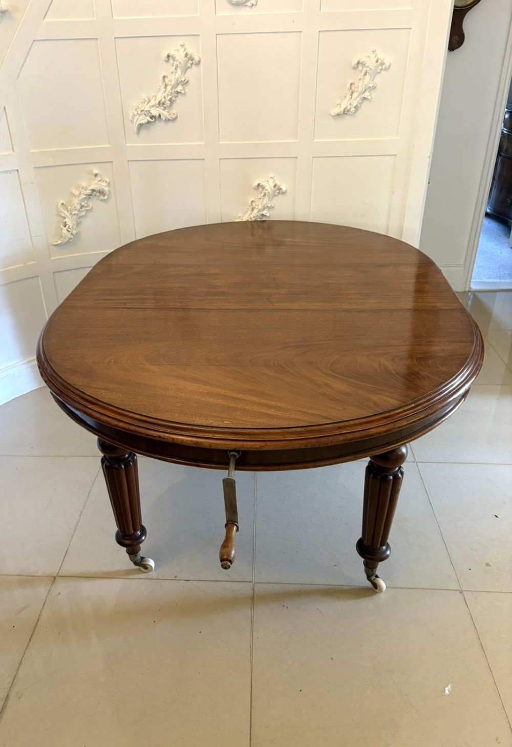 An antique Victorian oval mahogany dining table featuring a wind-out extending mechanism, fluted turned legs, and ceramic castors. View XII