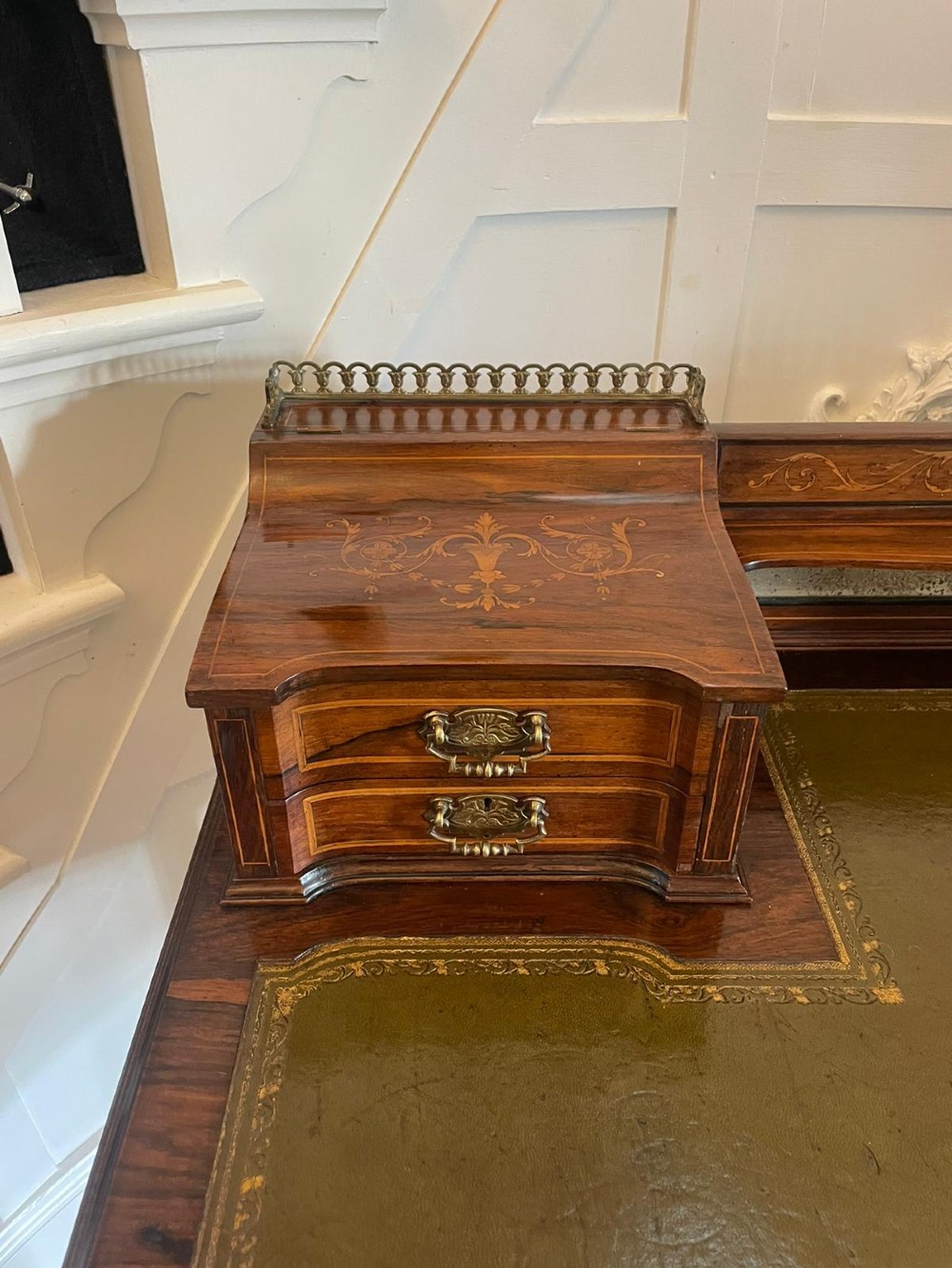 An antique Victorian inlaid writing desk with a green leather top, brass gallery railing, and multiple drawers on tapered legs with castors. View IV
