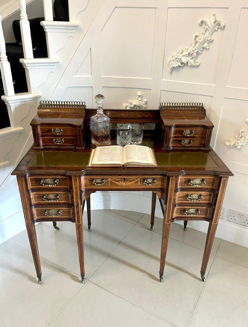 An antique Victorian inlaid writing desk with a green leather top, brass gallery railing, and multiple drawers on tapered legs with castors. View IX