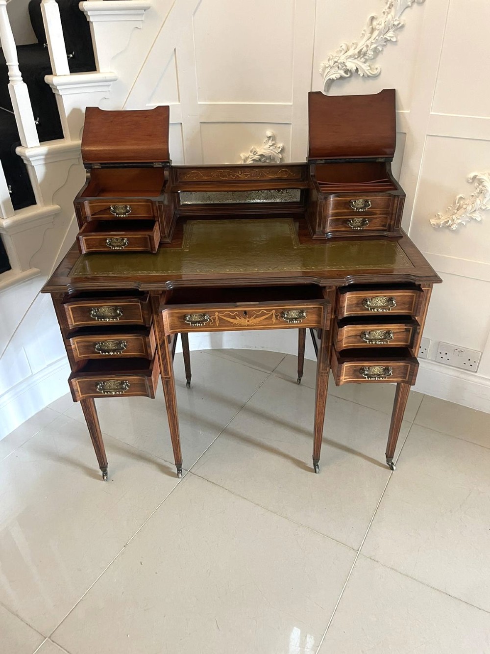 An antique Victorian inlaid writing desk with a green leather top, brass gallery railing, and multiple drawers on tapered legs with castors. View XI