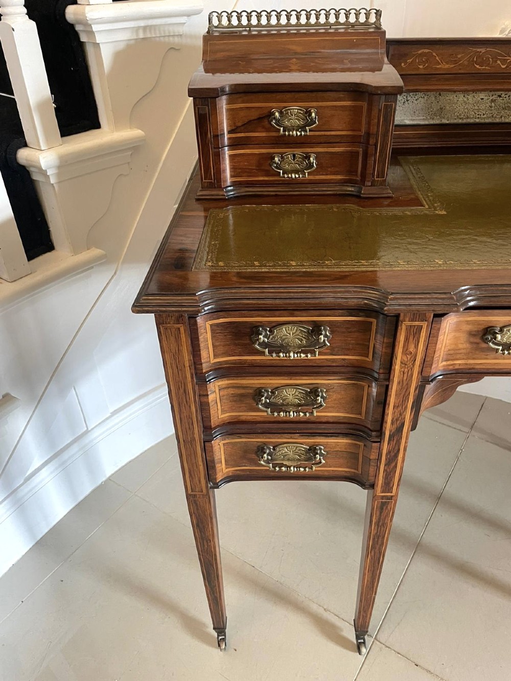 An antique Victorian inlaid writing desk with a green leather top, brass gallery railing, and multiple drawers on tapered legs with castors. View XII