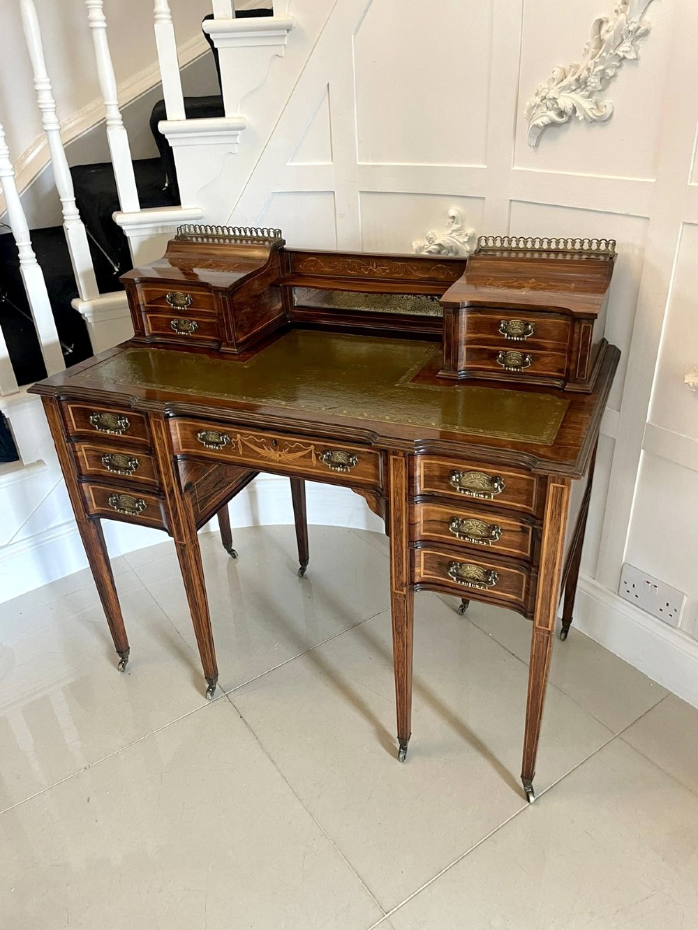 An antique Victorian inlaid writing desk with a green leather top, brass gallery railing, and multiple drawers on tapered legs with castors. View XIV