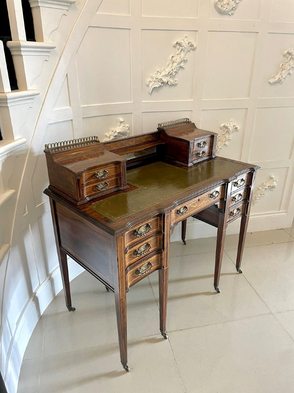 An antique Victorian inlaid writing desk with a green leather top, brass gallery railing, and multiple drawers on tapered legs with castors. View XV