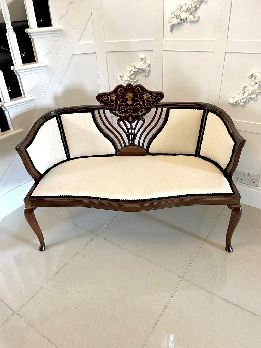 An antique Victorian mahogany settee featuring cream upholstered seating, curved arms, cabriole legs, and a central back splat with intricate floral wood inlay. View VI