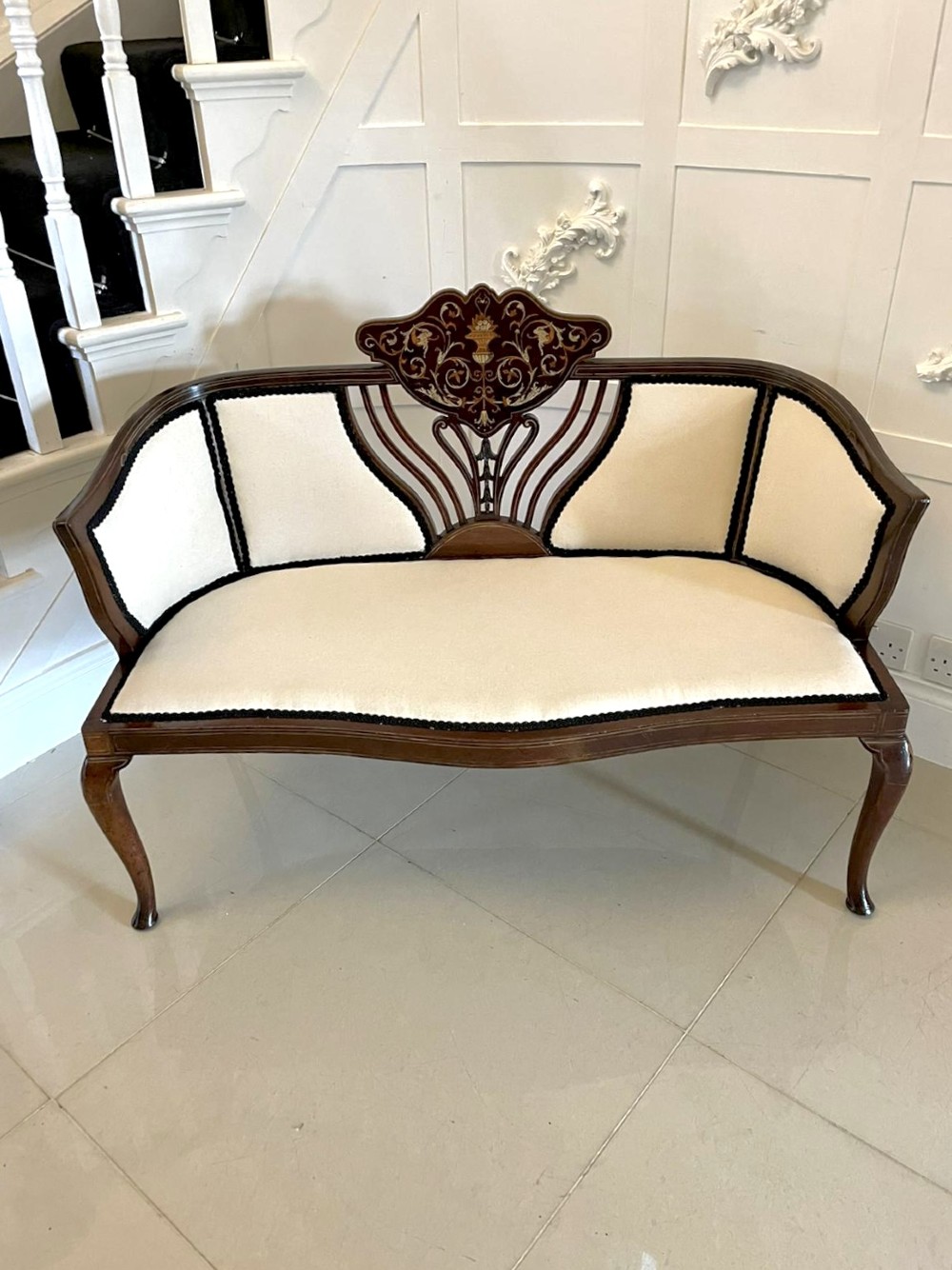 An antique Victorian mahogany settee featuring cream upholstered seating, curved arms, cabriole legs, and a central back splat with intricate floral wood inlay. View X
