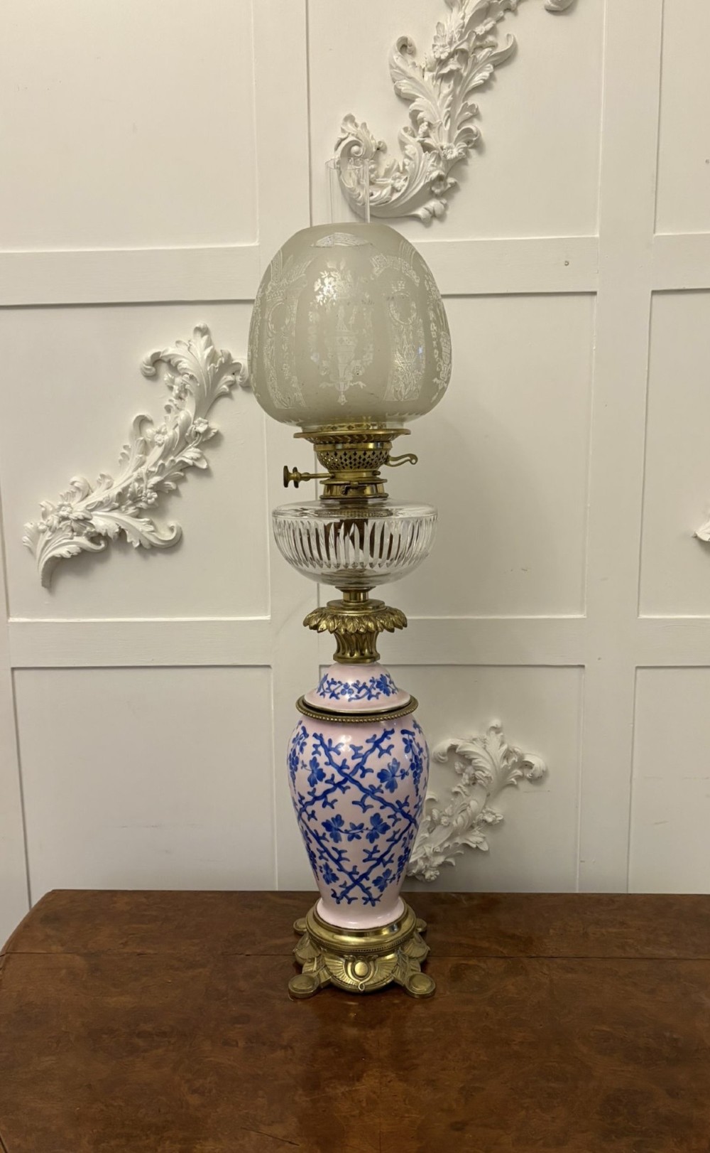 A tall Victorian oil lamp featuring a pink porcelain body with blue floral trellis decoration, a cast brass base, a cut glass reservoir, and an etched glass shade.