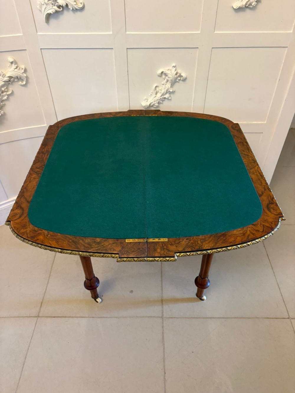 A Victorian burr walnut fold-over card table featuring geometric inlay, turned support legs on castors, and decorative brass edge moulding. View V