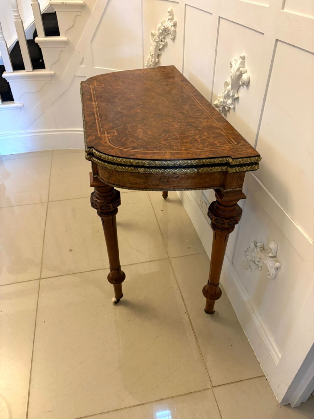 A Victorian burr walnut fold-over card table featuring geometric inlay, turned support legs on castors, and decorative brass edge moulding. View VII