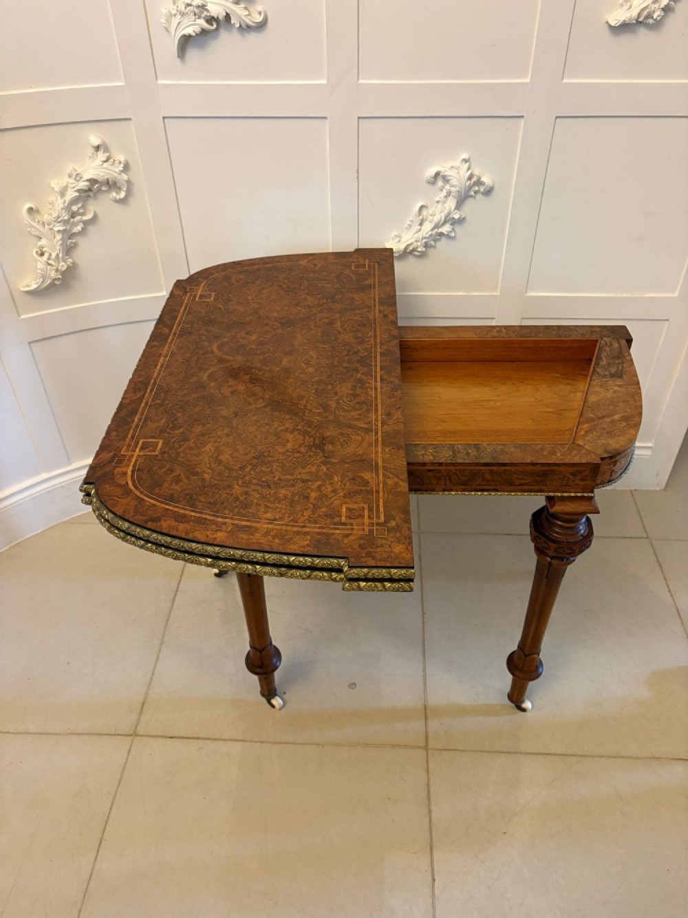 A Victorian burr walnut fold-over card table featuring geometric inlay, turned support legs on castors, and decorative brass edge moulding. View VIII