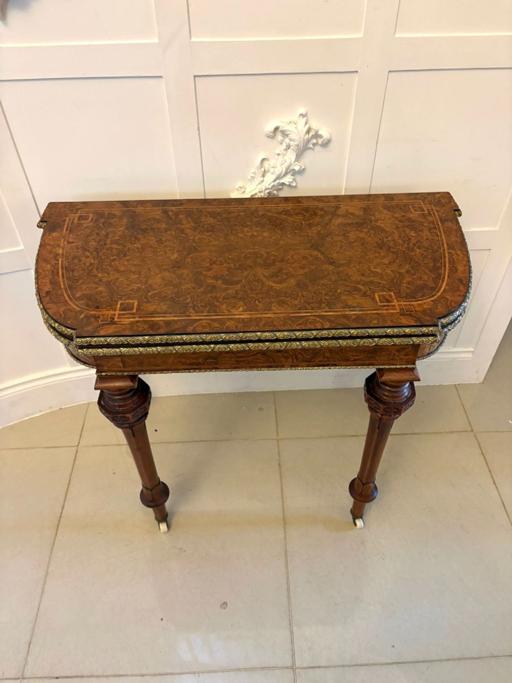 A Victorian burr walnut fold-over card table featuring geometric inlay, turned support legs on castors, and decorative brass edge moulding. View X
