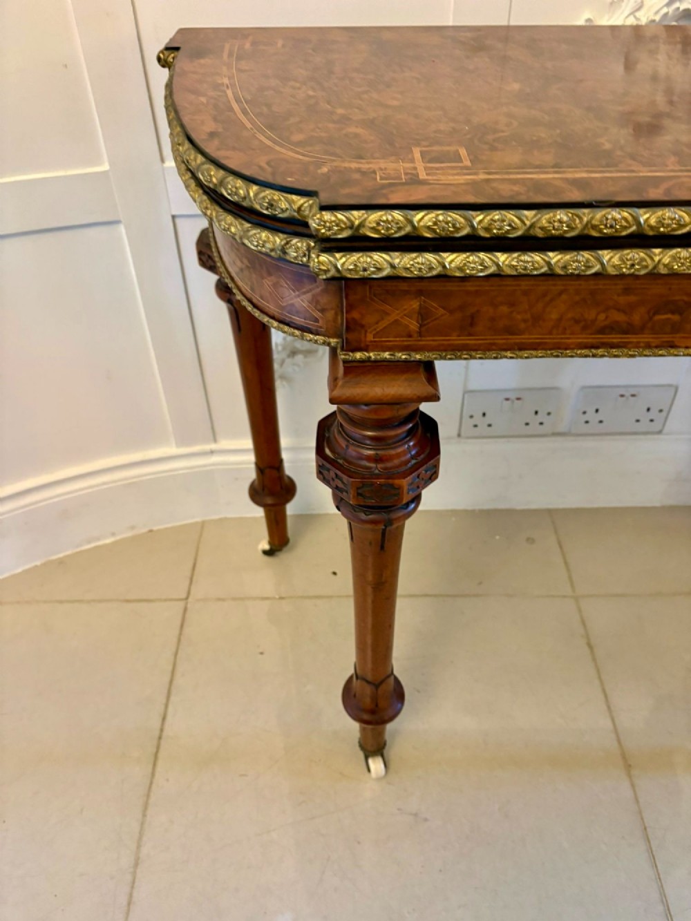 A Victorian burr walnut fold-over card table featuring geometric inlay, turned support legs on castors, and decorative brass edge moulding. View XII