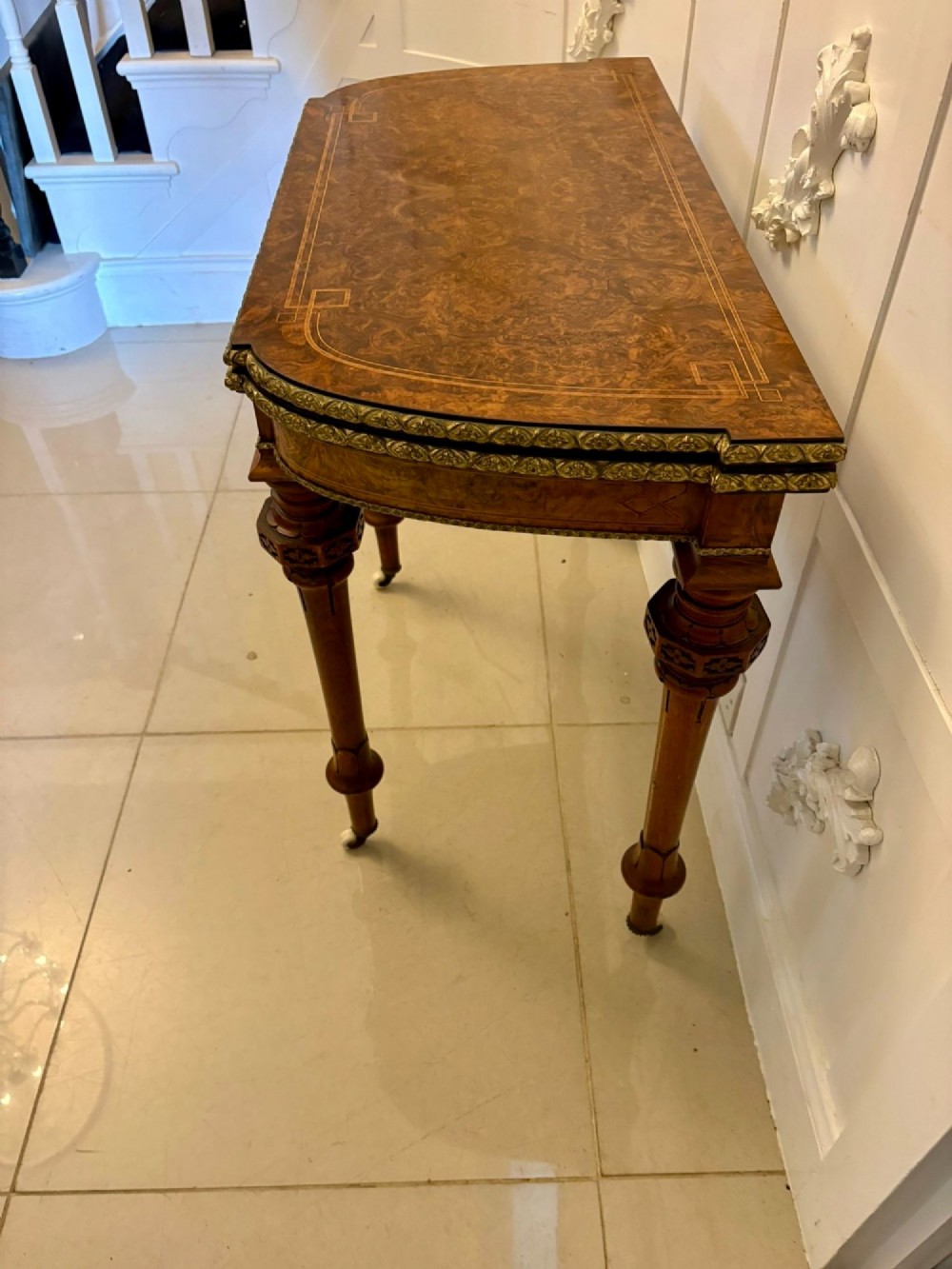 A Victorian burr walnut fold-over card table featuring geometric inlay, turned support legs on castors, and decorative brass edge moulding. View XIII