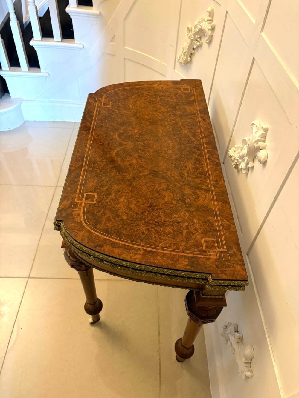 A Victorian burr walnut fold-over card table featuring geometric inlay, turned support legs on castors, and decorative brass edge moulding. View XIV