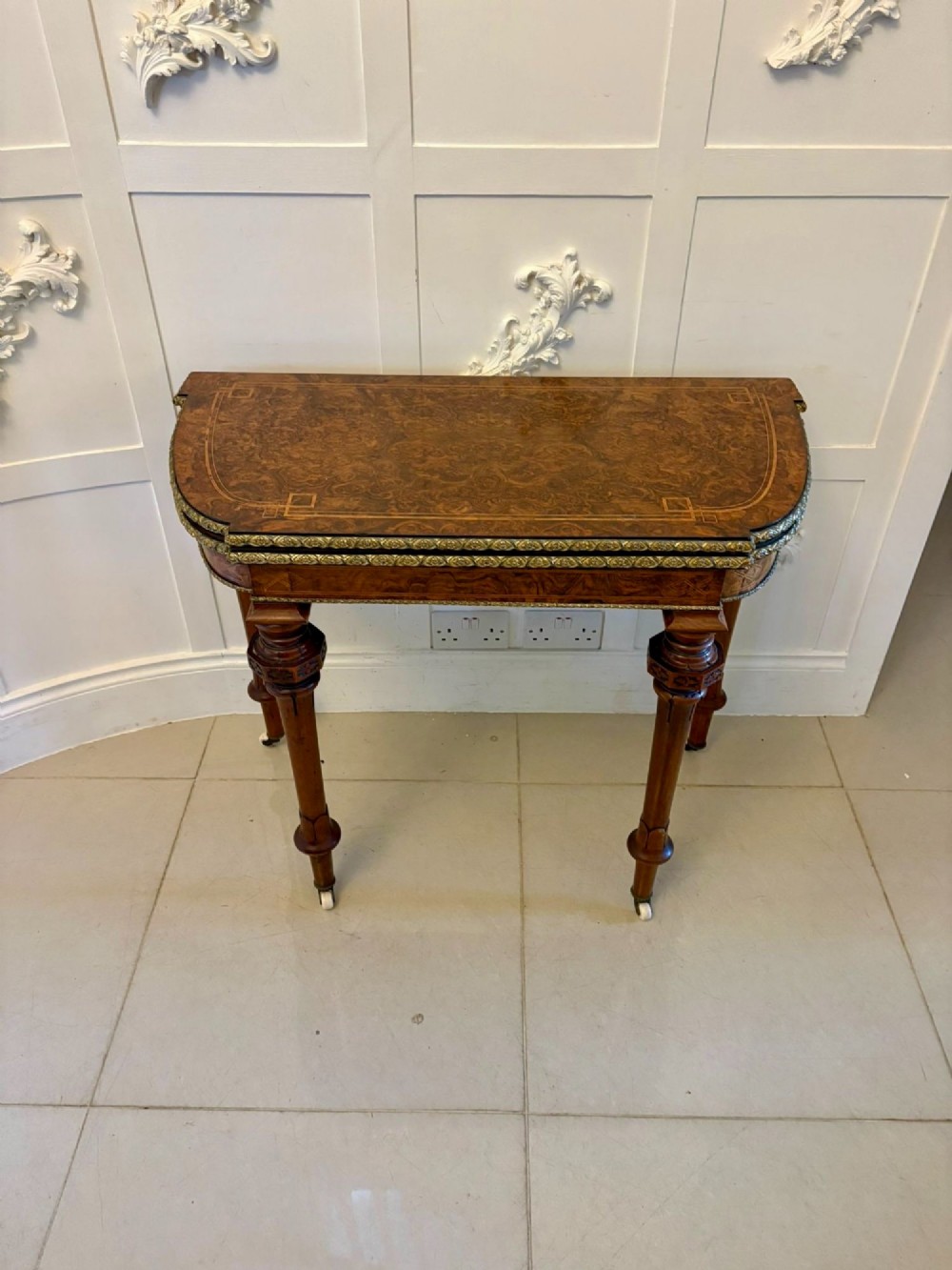 A Victorian burr walnut fold-over card table featuring geometric inlay, turned support legs on castors, and decorative brass edge moulding. View XV