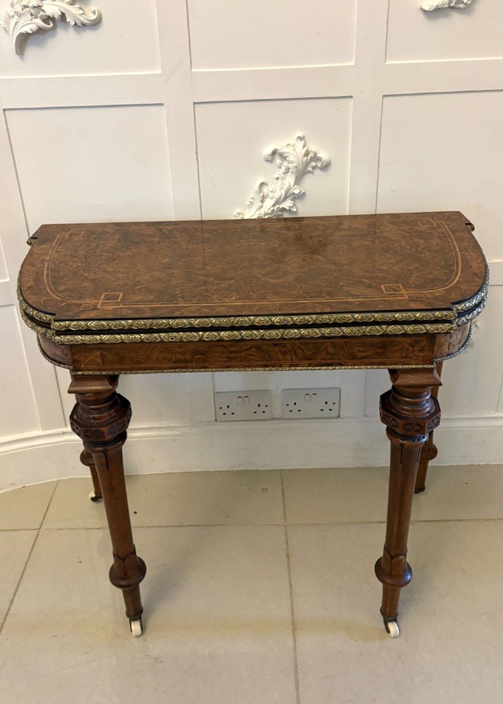 A Victorian burr walnut fold-over card table featuring geometric inlay, turned support legs on castors, and decorative brass edge moulding.