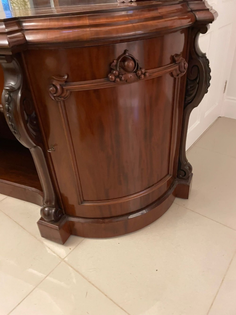 A Victorian carved mahogany sideboard featuring a curved front, open central bay, flanked cupboards, and a large arched mirror backrest adorned with scroll carving. View VII