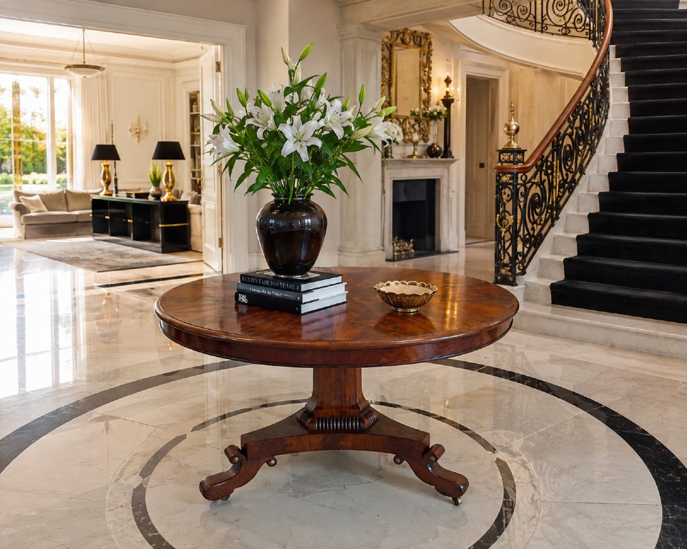 A circular Regency tilt-top centre table featuring highly figured flame mahogany veneers, shown in the upright position on a scrolled pedestal base with brass castors. View II