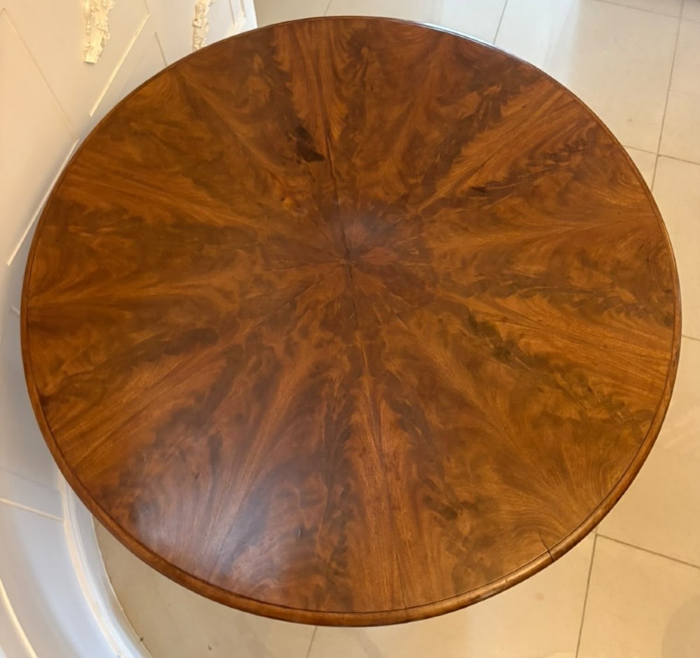 A circular Regency tilt-top centre table featuring highly figured flame mahogany veneers, shown in the upright position on a scrolled pedestal base with brass castors. View V
