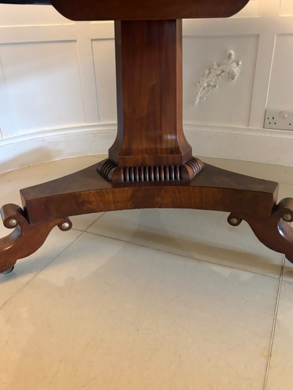 A circular Regency tilt-top centre table featuring highly figured flame mahogany veneers, shown in the upright position on a scrolled pedestal base with brass castors. View VI