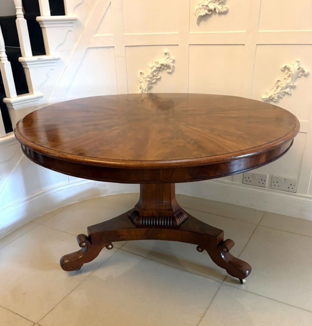 A circular Regency tilt-top centre table featuring highly figured flame mahogany veneers, shown in the upright position on a scrolled pedestal base with brass castors. View XII