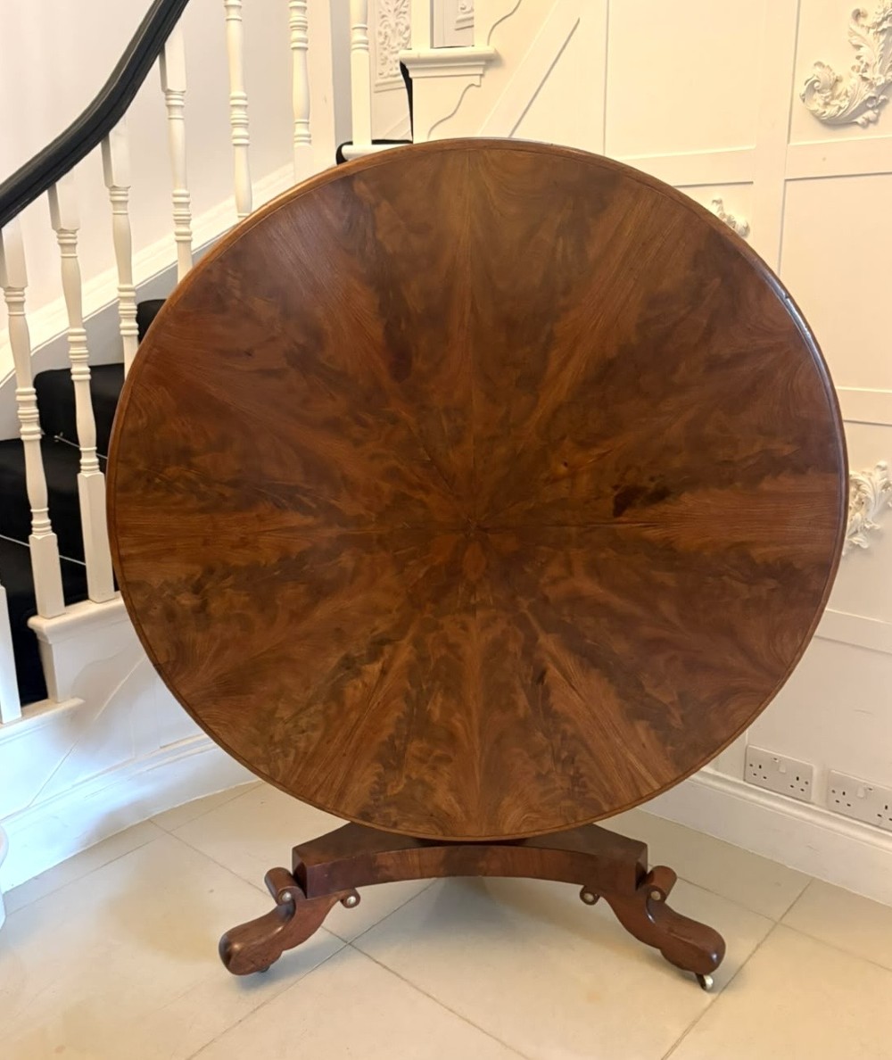 A circular Regency tilt-top centre table featuring highly figured flame mahogany veneers, shown in the upright position on a scrolled pedestal base with brass castors.