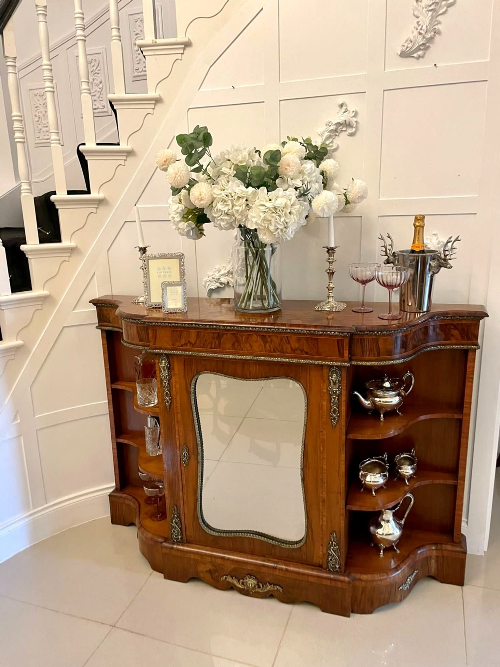 A Victorian burr walnut credenza featuring a central glass door, open serpentine display shelves, and detailed brass ormolu mounts. View VI