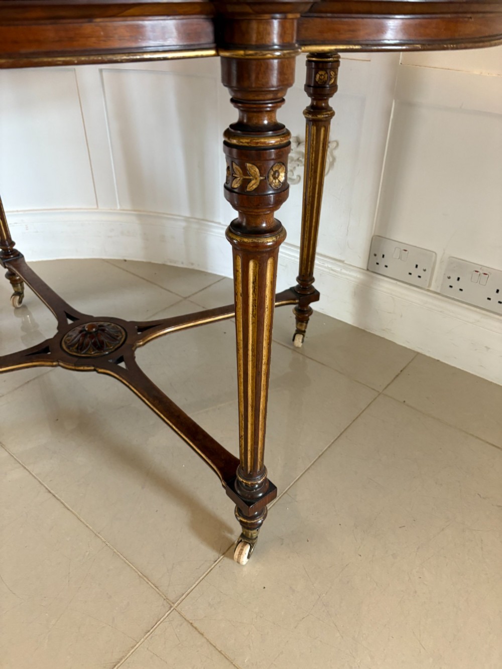 An antique Victorian burr walnut centre table featuring a shaped top with marquetry inlay, turned fluted legs, an X-frame stretcher, and ceramic castors. View VI