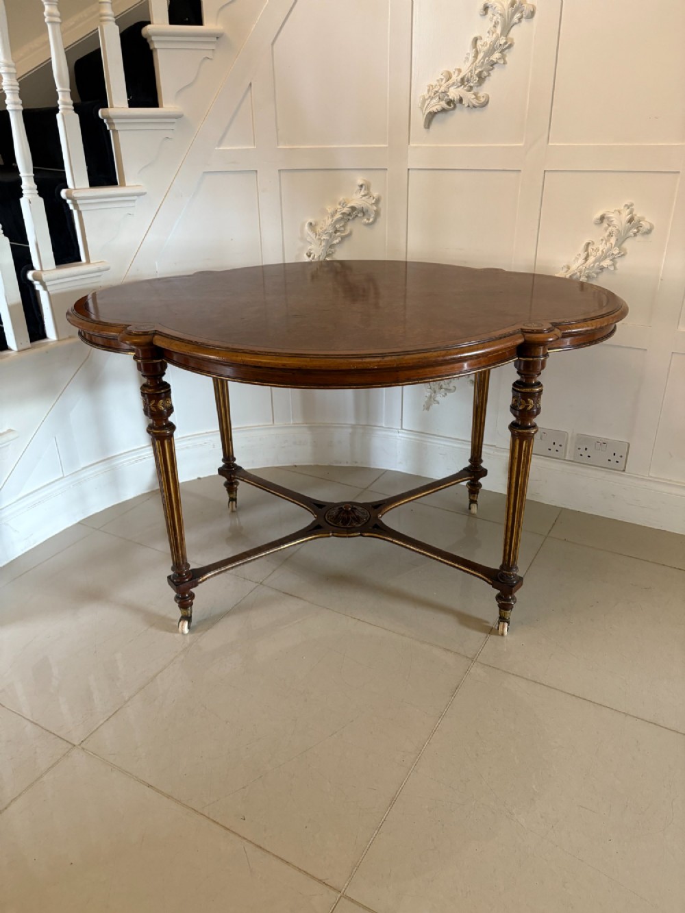 An antique Victorian burr walnut centre table featuring a shaped top with marquetry inlay, turned fluted legs, an X-frame stretcher, and ceramic castors. View XII