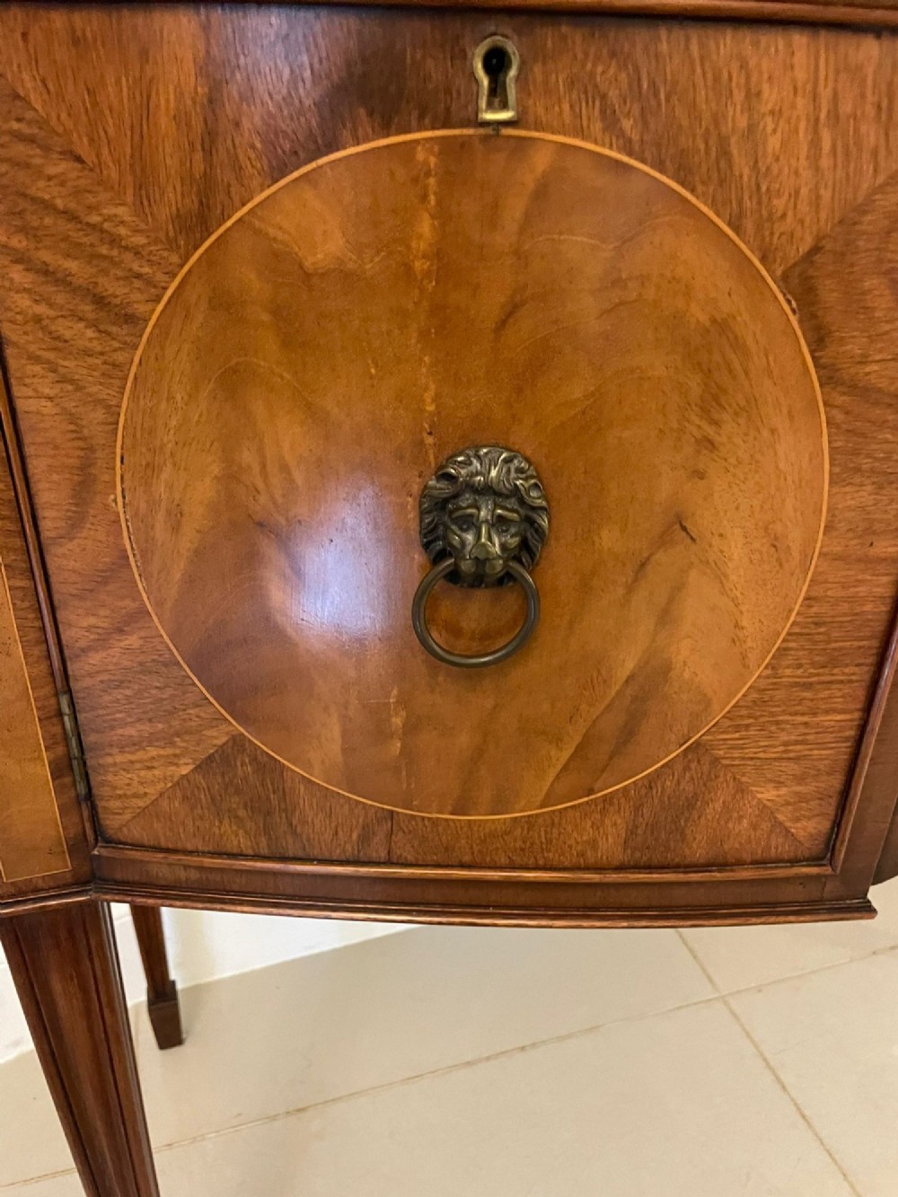 An antique bow-fronted mahogany sideboard featuring a central drawer, two side cupboards with oval inlaid panels, brass lion-mask ring handles, and square tapered legs. View III