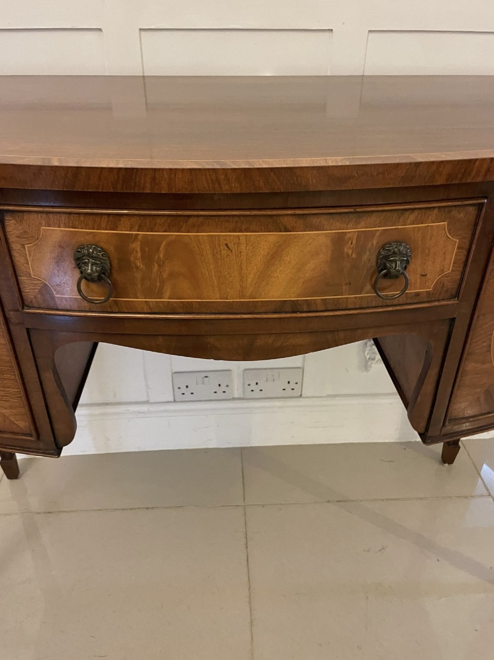 An antique bow-fronted mahogany sideboard featuring a central drawer, two side cupboards with oval inlaid panels, brass lion-mask ring handles, and square tapered legs. View IV