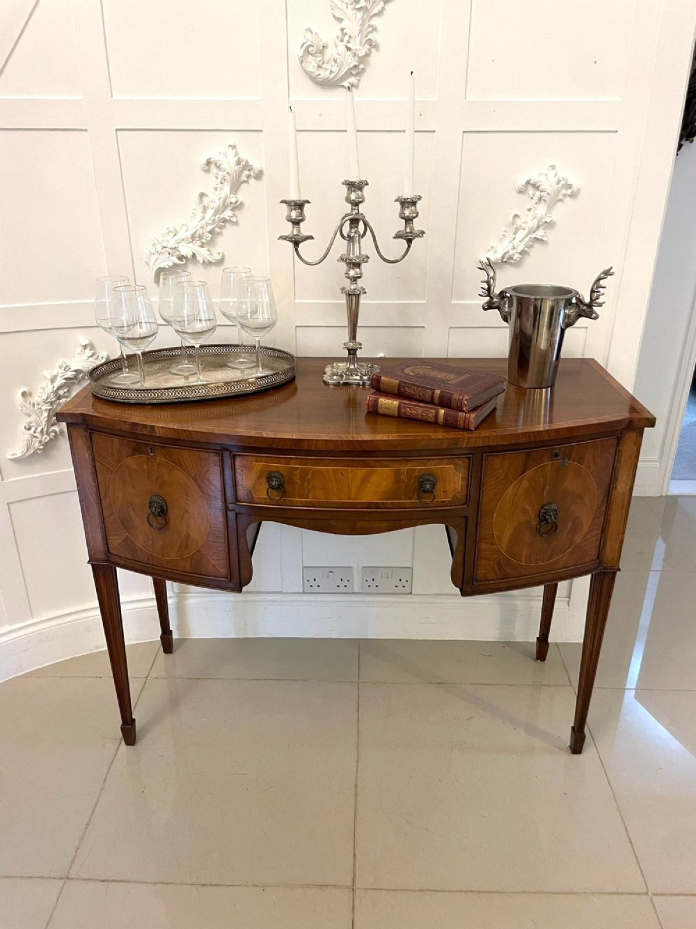 An antique bow-fronted mahogany sideboard featuring a central drawer, two side cupboards with oval inlaid panels, brass lion-mask ring handles, and square tapered legs. View V
