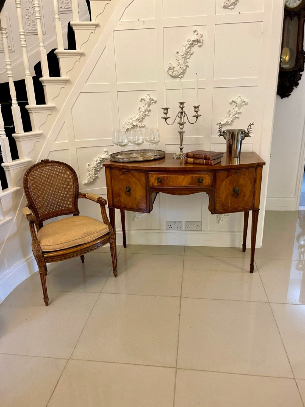 An antique bow-fronted mahogany sideboard featuring a central drawer, two side cupboards with oval inlaid panels, brass lion-mask ring handles, and square tapered legs. View VI