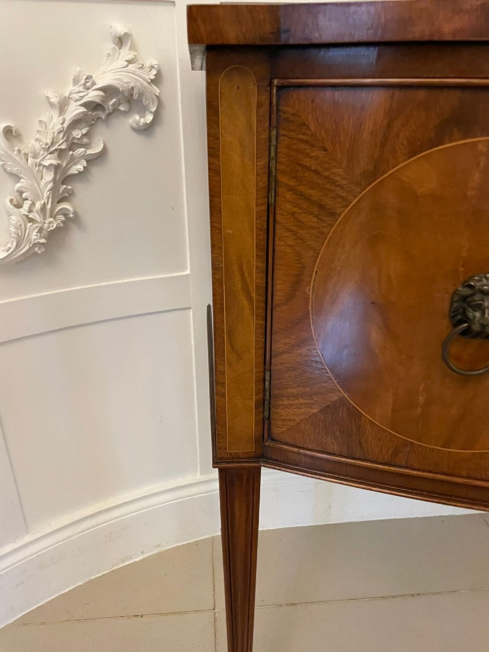 An antique bow-fronted mahogany sideboard featuring a central drawer, two side cupboards with oval inlaid panels, brass lion-mask ring handles, and square tapered legs. View VII
