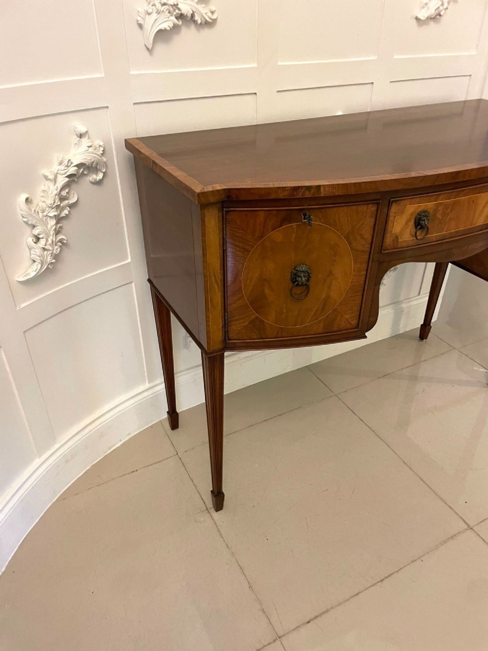An antique bow-fronted mahogany sideboard featuring a central drawer, two side cupboards with oval inlaid panels, brass lion-mask ring handles, and square tapered legs. View VIII