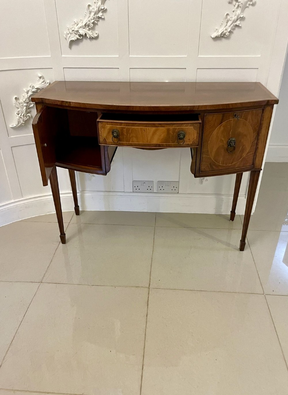 An antique bow-fronted mahogany sideboard featuring a central drawer, two side cupboards with oval inlaid panels, brass lion-mask ring handles, and square tapered legs. View IX