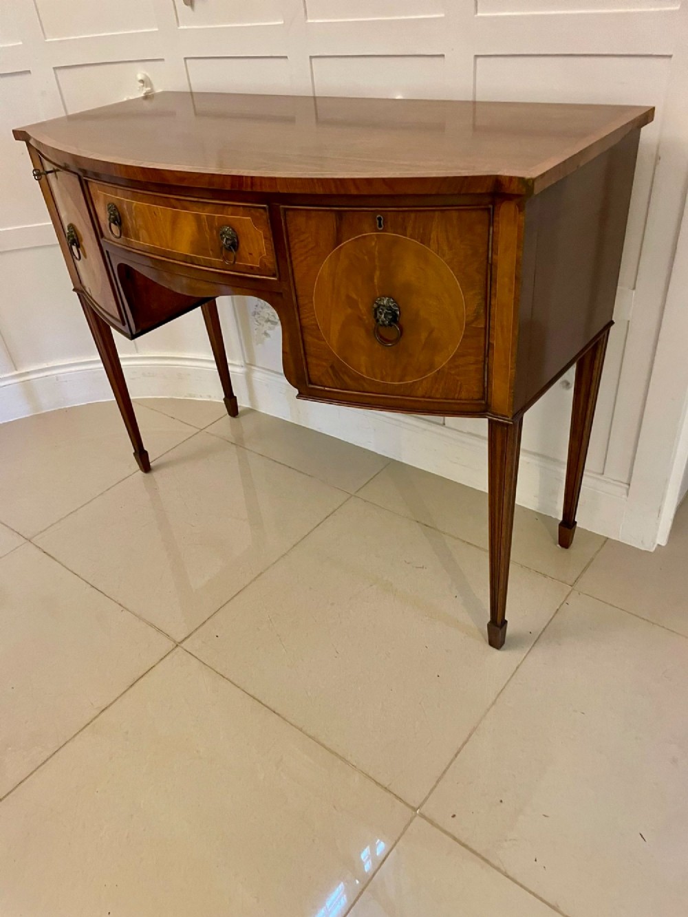 An antique bow-fronted mahogany sideboard featuring a central drawer, two side cupboards with oval inlaid panels, brass lion-mask ring handles, and square tapered legs. View X