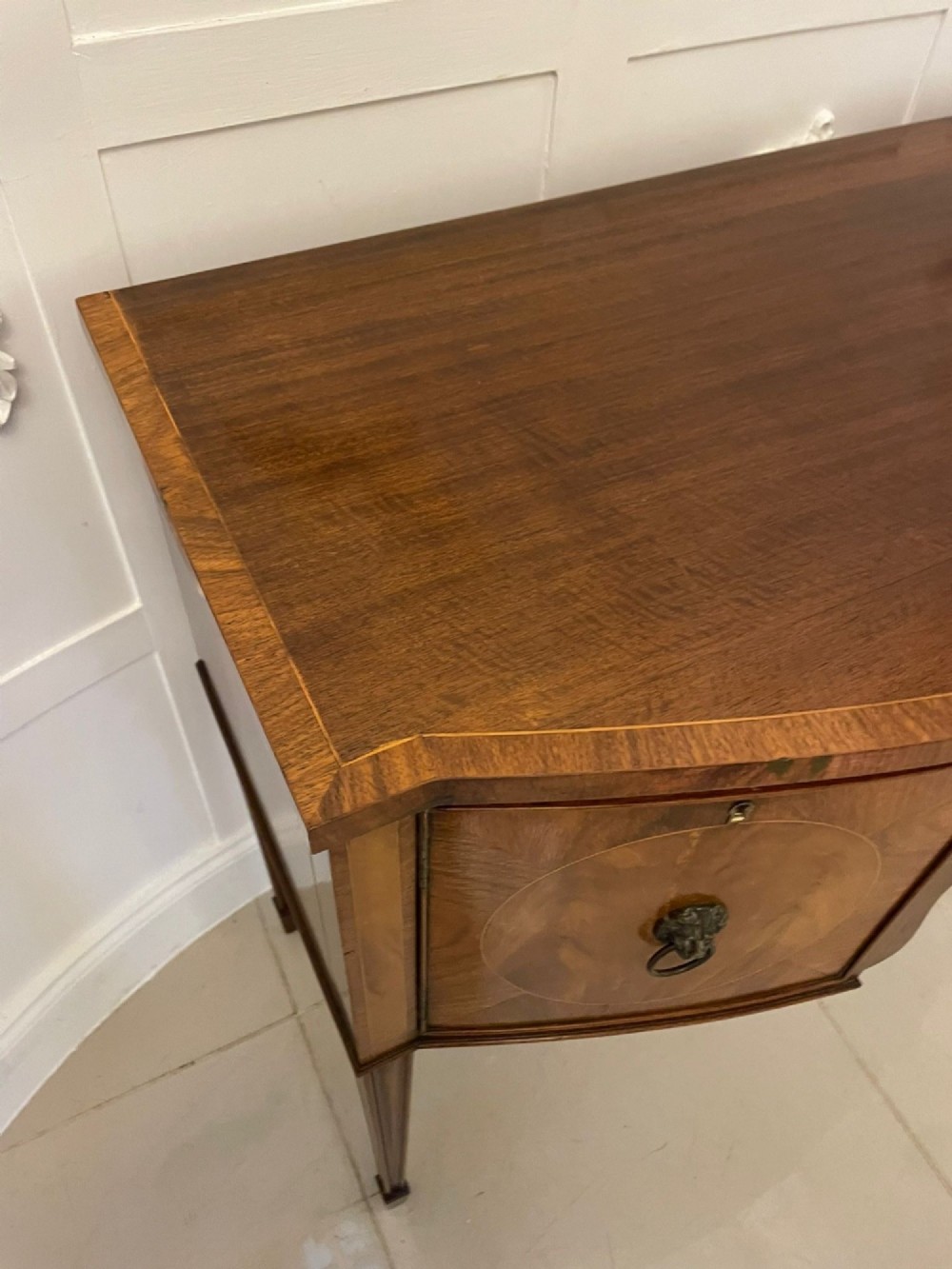 An antique bow-fronted mahogany sideboard featuring a central drawer, two side cupboards with oval inlaid panels, brass lion-mask ring handles, and square tapered legs. View XII