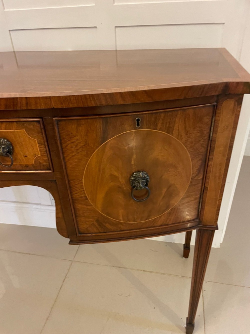 An antique bow-fronted mahogany sideboard featuring a central drawer, two side cupboards with oval inlaid panels, brass lion-mask ring handles, and square tapered legs. View XIII