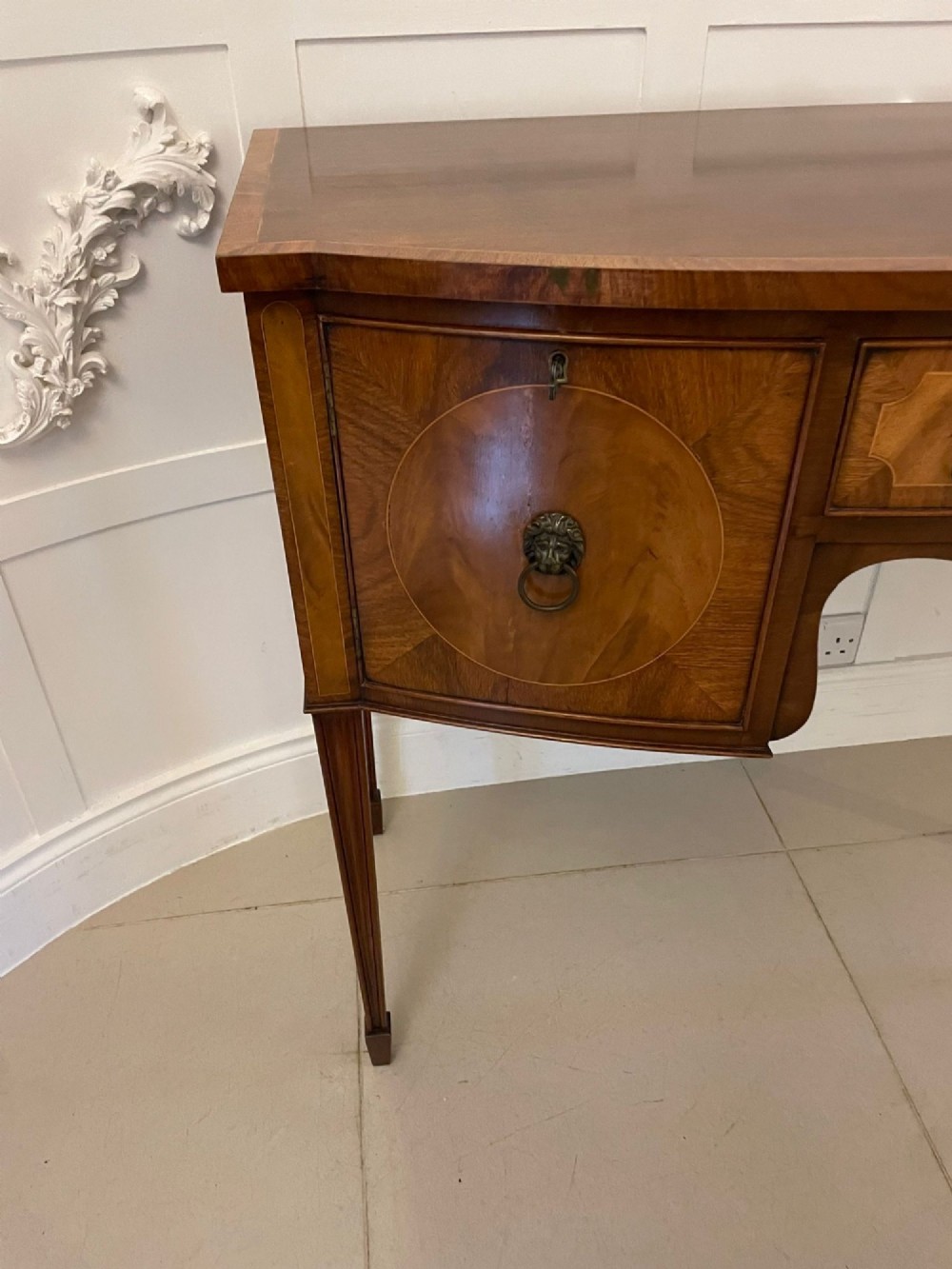 An antique bow-fronted mahogany sideboard featuring a central drawer, two side cupboards with oval inlaid panels, brass lion-mask ring handles, and square tapered legs. View XIV