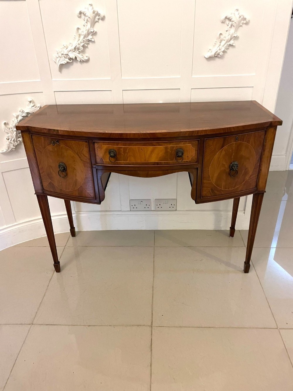 An antique bow-fronted mahogany sideboard featuring a central drawer, two side cupboards with oval inlaid panels, brass lion-mask ring handles, and square tapered legs.