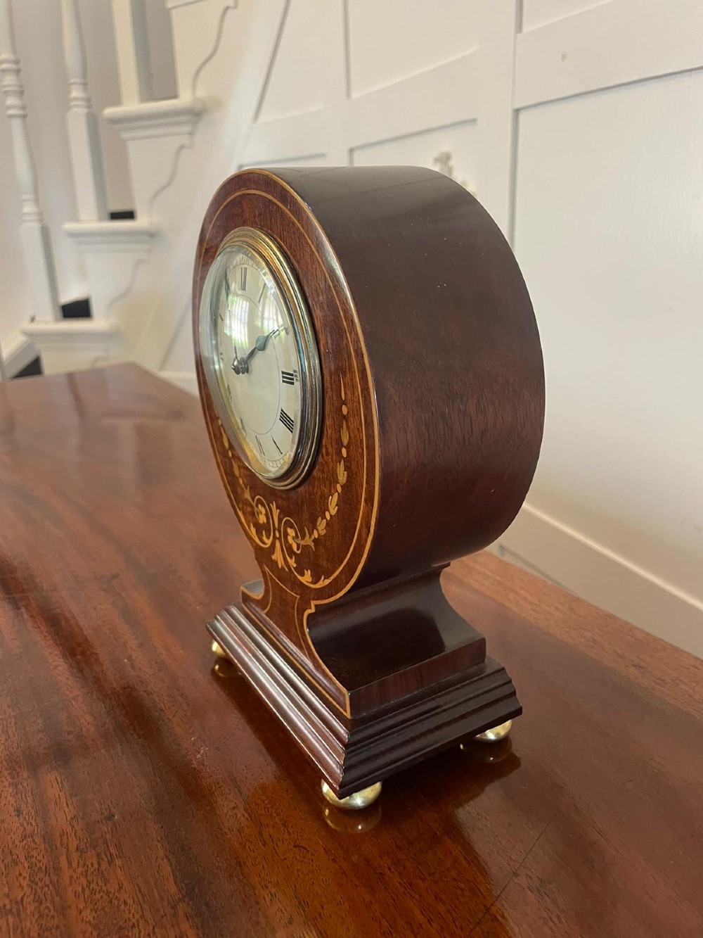 An Edwardian mahogany mantle clock featuring floral satinwood inlay, a white dial with Roman numerals, a brass bezel, and small brass bun feet. View IV