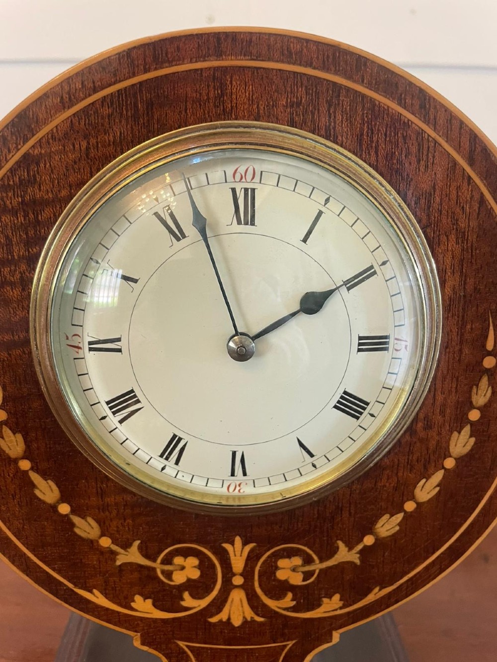 An Edwardian mahogany mantle clock featuring floral satinwood inlay, a white dial with Roman numerals, a brass bezel, and small brass bun feet. View V