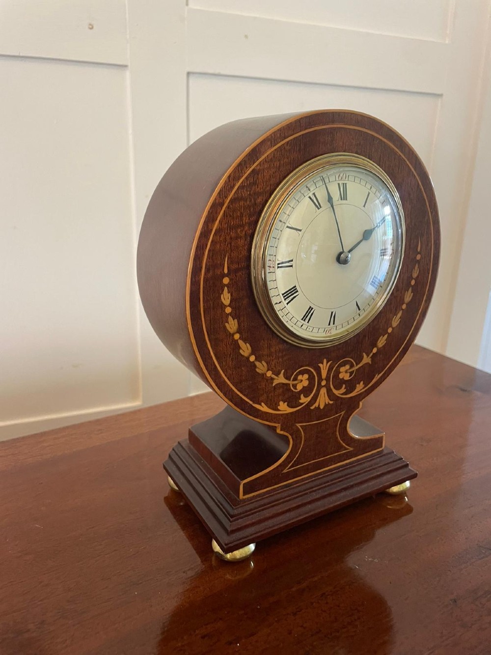 An Edwardian mahogany mantle clock featuring floral satinwood inlay, a white dial with Roman numerals, a brass bezel, and small brass bun feet. View VI