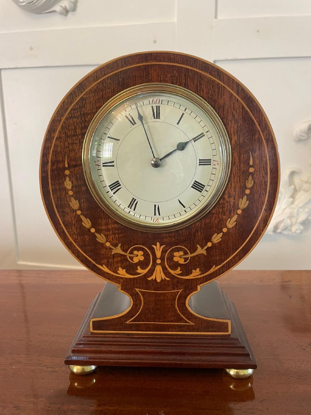 An Edwardian mahogany mantle clock featuring floral satinwood inlay, a white dial with Roman numerals, a brass bezel, and small brass bun feet.