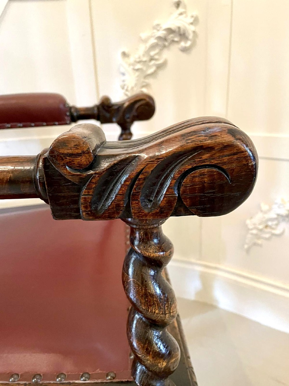 A high-backed Victorian carved oak armchair featuring barley twist supports, acanthus leaf carvings, and a burgundy leather seat with brass stud detail. View VI