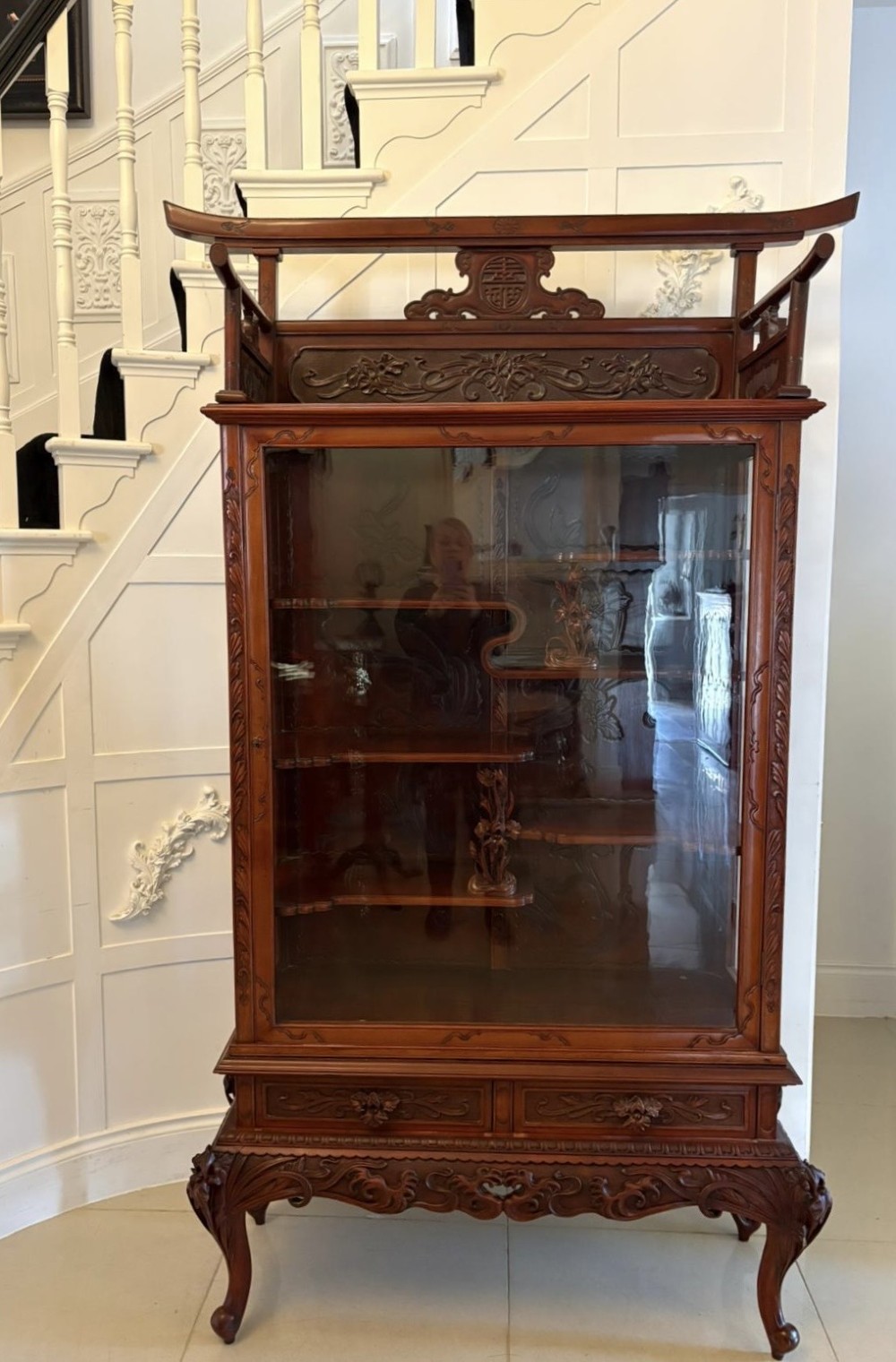 A carved reddish-brown hardwood display cabinet featuring a pagoda cresting, glass door, asymmetrical interior shelves, lower drawers, and cabriole legs.