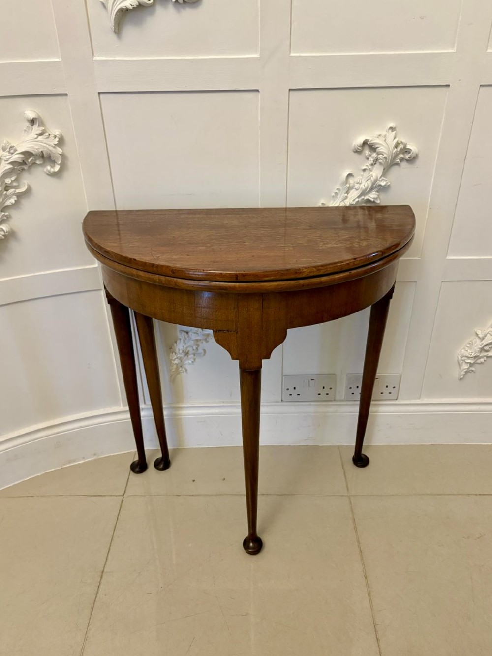 An antique warm brown mahogany demi-lune card table featuring tapered legs with pad feet and a fold-over top.
