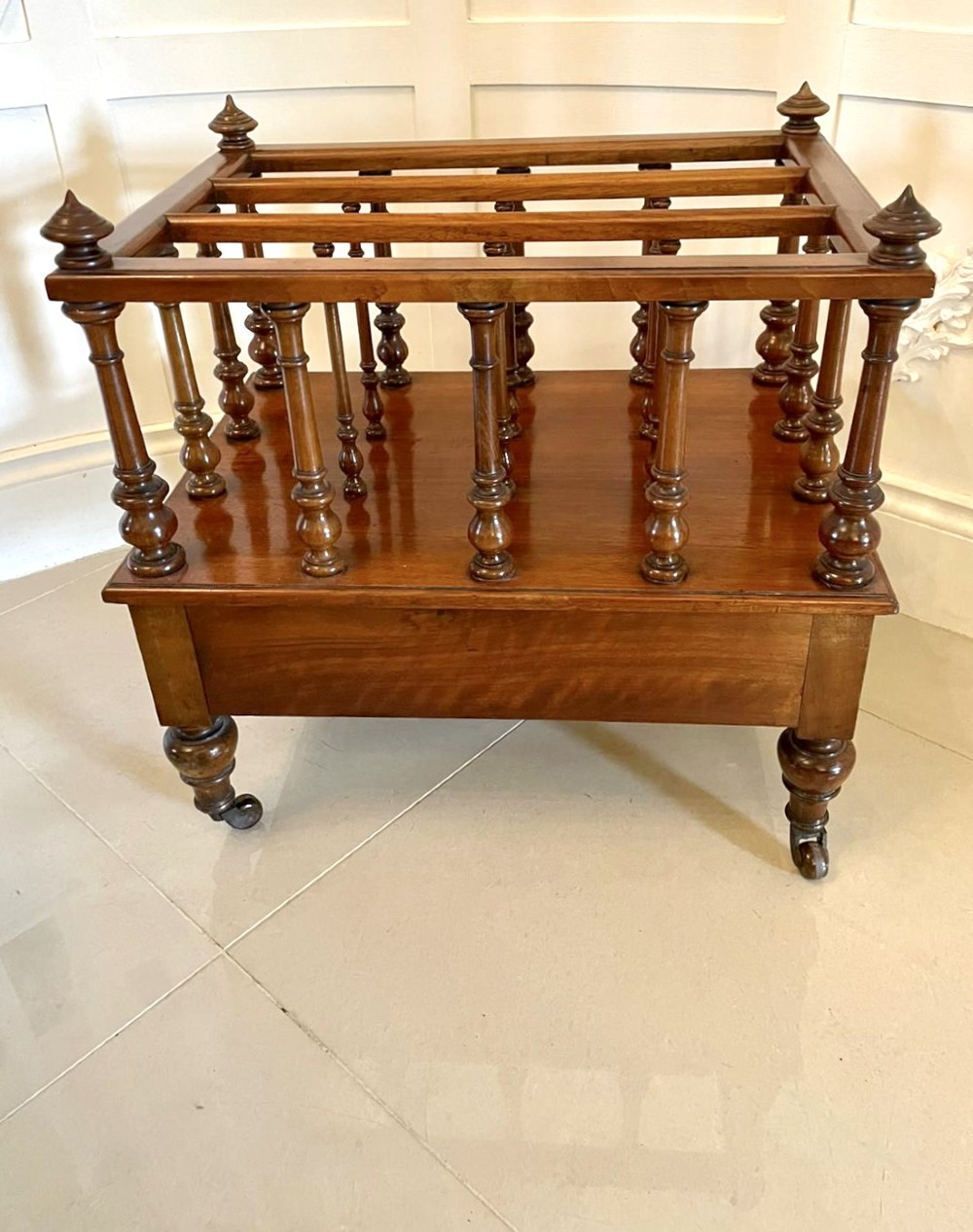 A Victorian figured walnut Canterbury featuring multiple vertical turned spindles, corner finials, a lower base drawer with two knobs, and brass castors. View VI