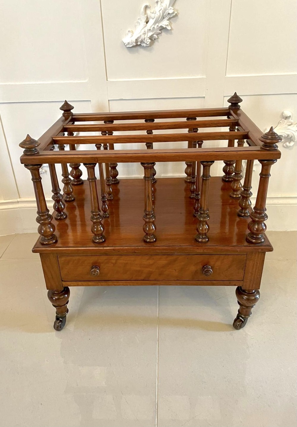 A Victorian figured walnut Canterbury featuring multiple vertical turned spindles, corner finials, a lower base drawer with two knobs, and brass castors. View X