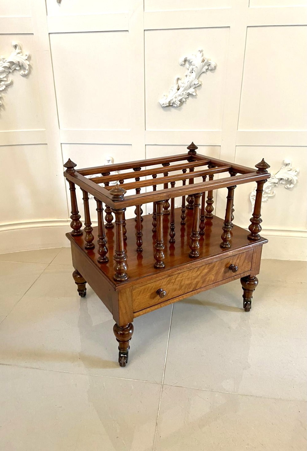 A Victorian figured walnut Canterbury featuring multiple vertical turned spindles, corner finials, a lower base drawer with two knobs, and brass castors.